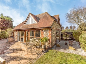 A stone house with a tiled roof, a patio with a table and chairs, and a garden with gravel paths and plants.