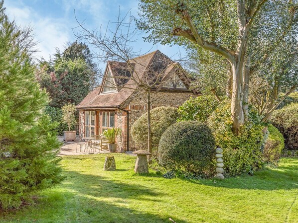 A stone house with a tiled roof, surrounded by a well-maintained garden with a lawn, a wooden bench, and a stone pathway leading to the entrance.