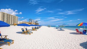 A beach with rows of sun loungers and umbrellas, a hotel building, and clear blue skies.