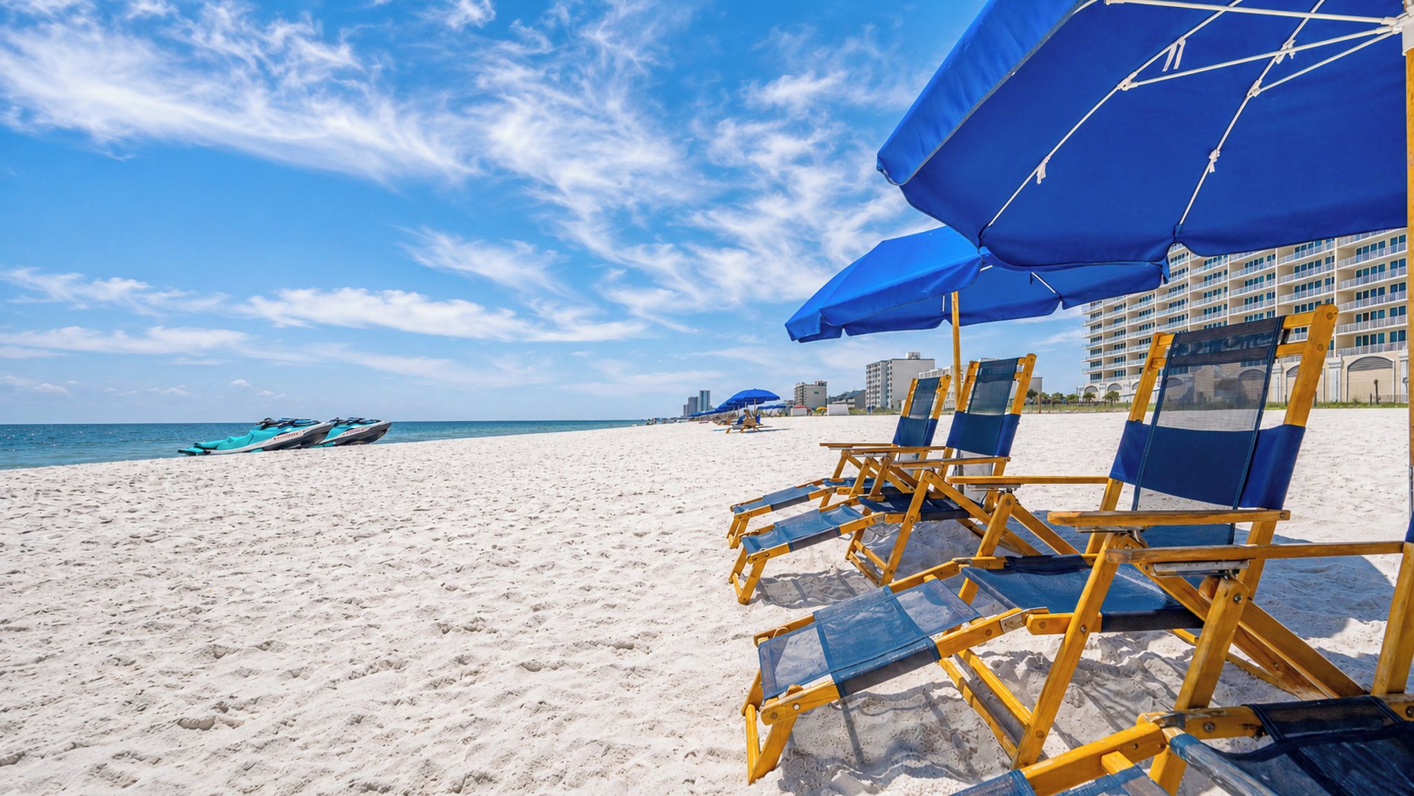 A beach with rows of beach chairs and umbrellas, a clear sky, and a distant view of a hotel and boats.
