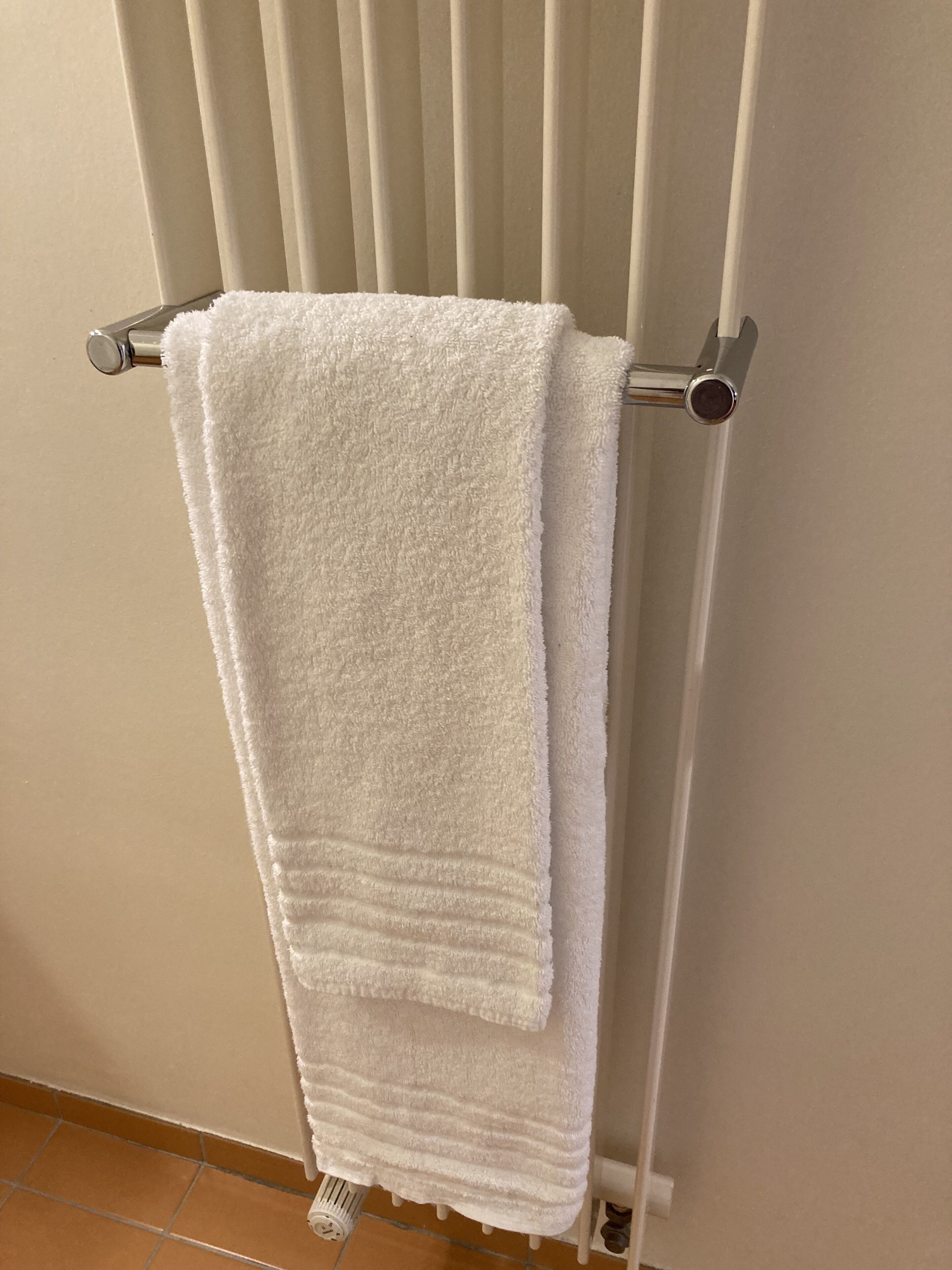 A white towel hanging on a metal towel rack in a bathroom with a tiled floor.