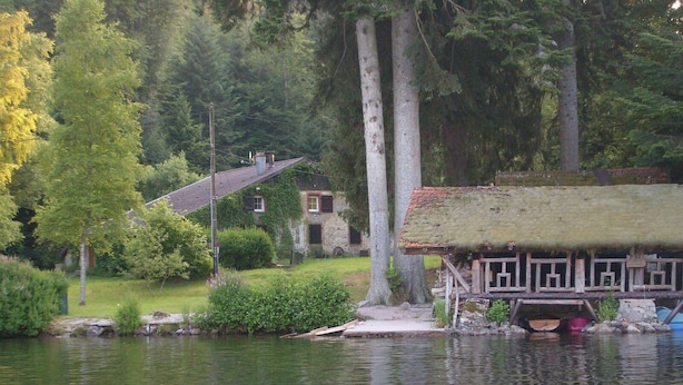 Una cabaña junto al lago con techo de paja, rodeada por un bosque frondoso y un pequeño muelle.