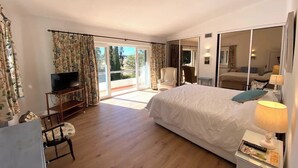 Une chambre à coucher avec un grand lit, un téléviseur posé sur un meuble en bois, une chaise et une table avec une lampe.