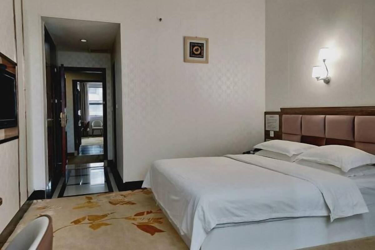 Habitación de hotel con cama, mesita de noche, lámpara de pared y puerta que da a un pasillo.