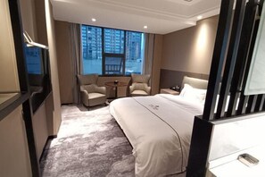 A hotel room with a large bed, a small table, two armchairs, and a view of buildings through the window.