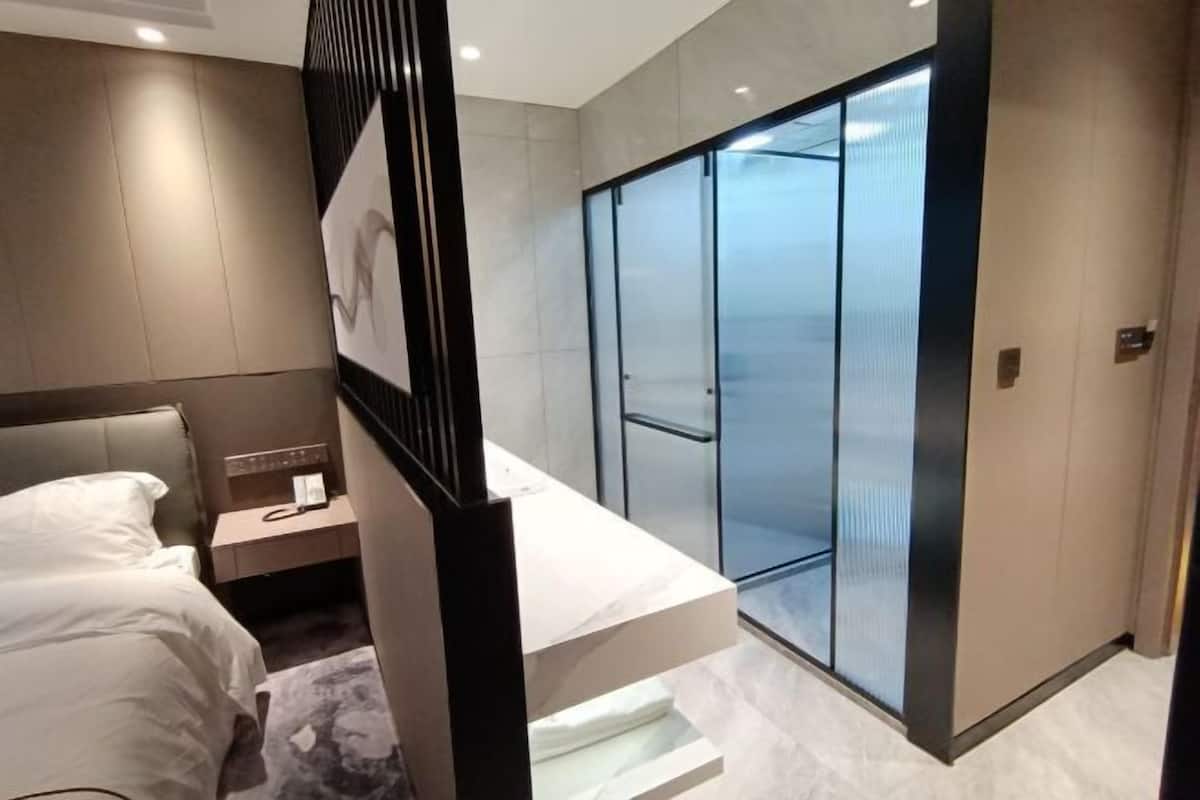 Una habitación de hotel moderna con una cama, una mesita de noche, un teléfono y la puerta de un ascensor de vidrio.