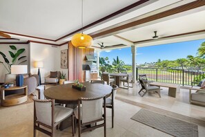 A spacious living area with a round wooden table, comfortable seating, a ceiling fan, and a view of the outdoors.