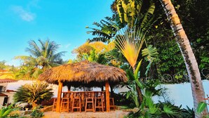 A thatched-roof hut with wooden benches, surrounded by tropical plants and palm trees. - Exclusive Luxury Getaway: 6 BRM Tropical Villa, Just 5 Minutes from Beaches (Sosúa)