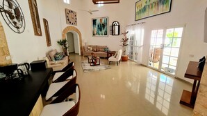 A spacious living area with a sofa set, a coffee table, and a dining area with chairs and a table. There is a large window with curtains, a clock on the wall, and a painting above the sofa. - Exclusive Luxury Getaway: 6 BRM Tropical Villa, Just 5 Minutes from Beaches (Sosúa)