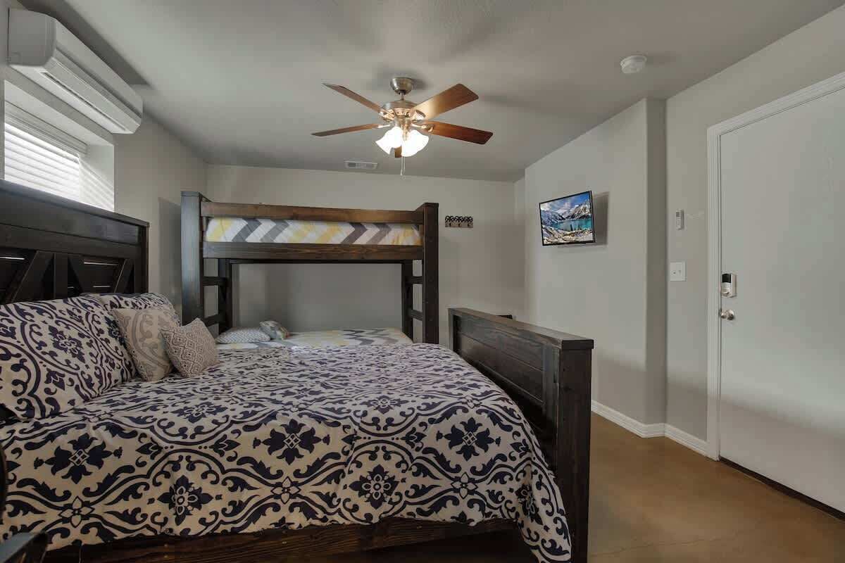 A bedroom with a bunk bed, a ceiling fan, a bed with patterned bedding, a bedside table, and a door.