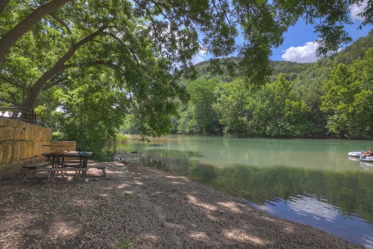 A riverside picnic area with tables and benches, surrounded by trees and a calm river.