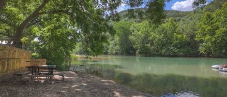 A riverside picnic area with tables and benches, surrounded by trees and a calm river.
