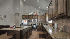 A spacious kitchen with dark wooden cabinets, a granite countertop, stainless steel appliances, and a large island with stools. - Easy Tubing!-Right on the River! (New Braunfels)