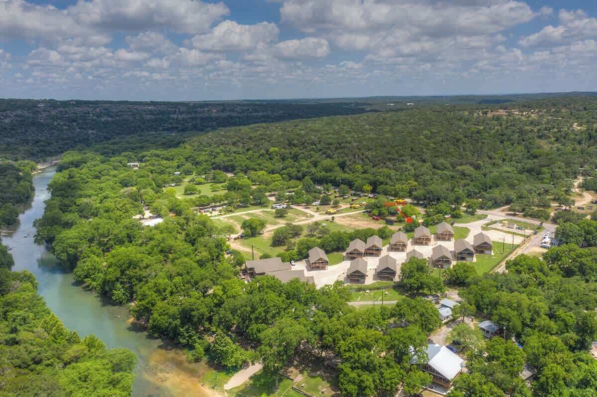 An aerial view of a resort with multiple cabins arranged in a semi-circular pattern around a central courtyard, with a river running through the property.