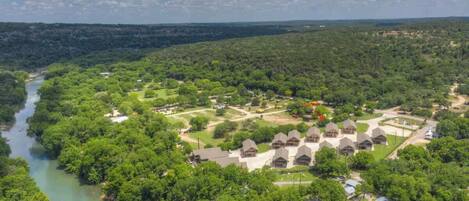 An aerial view of a resort with multiple cabins arranged in a semi-circular pattern around a central courtyard, with a river running through the property.
