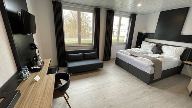 A modern hotel room with a bed, a desk, a chair, a TV, and a window with curtains. - Yebs Hotel (Guenzburg)