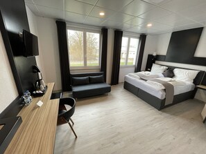 A modern hotel room with a bed, a desk, a chair, a TV, and a window with curtains.