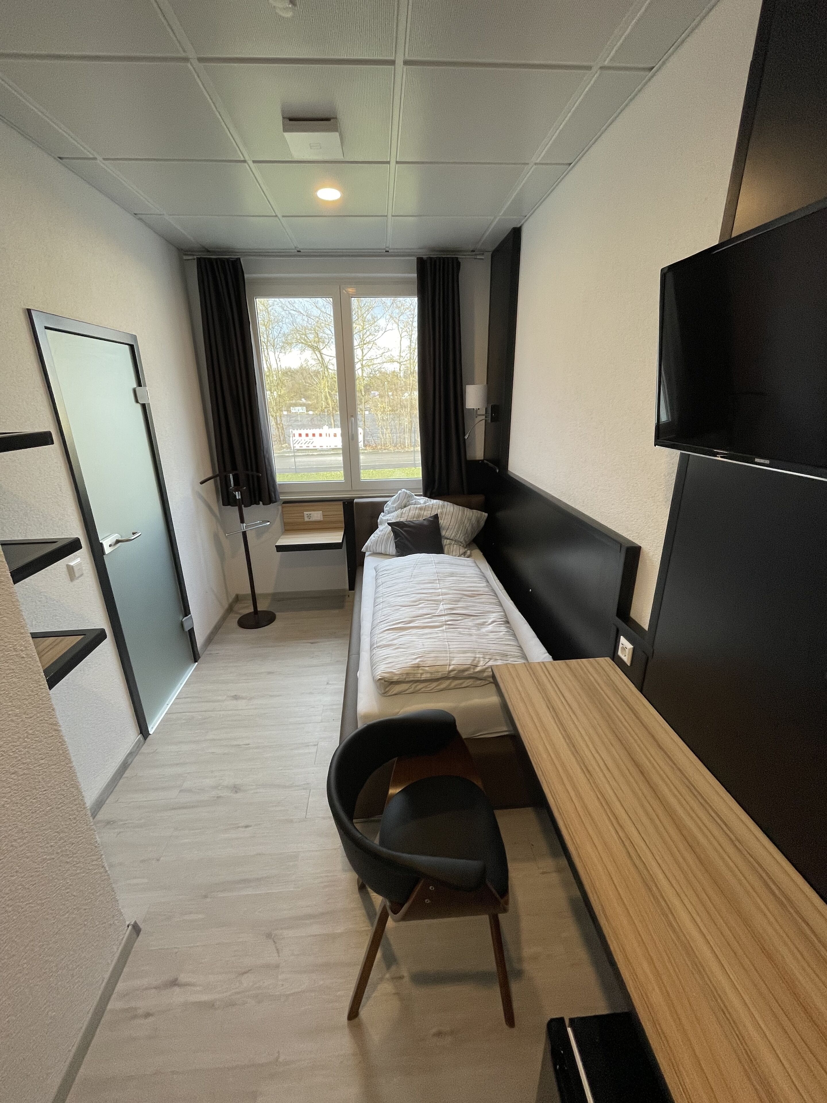 Habitación de hotel pequeña con una cama individual, un escritorio, una silla, un televisor y una ventana con cortinas.