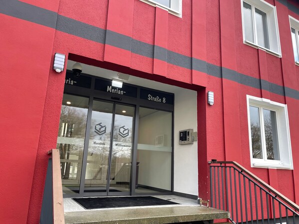 A building entrance with a red facade, glass doors, and a sign reading "Merian - Straße 8".