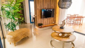 Ein modernes Wohnzimmer mit an einer Holzwand montiertem Fernseher, einem runden Holztisch, einer gewobenen Bank und einem Essbereich mit Holzkästen.
