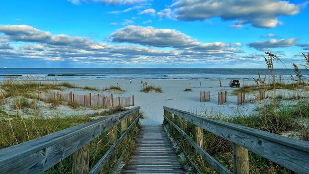 En träpromenad som leder till en strand med sanddyner och en klar himmel.