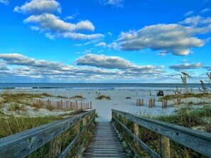 A wooden boardwalk leading to a beach with dunes and a clear sky.