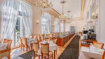 Un comedor amplio con varias mesas y sillas, candelabros elegantes y ventanas grandes con cortinas translúcidas.