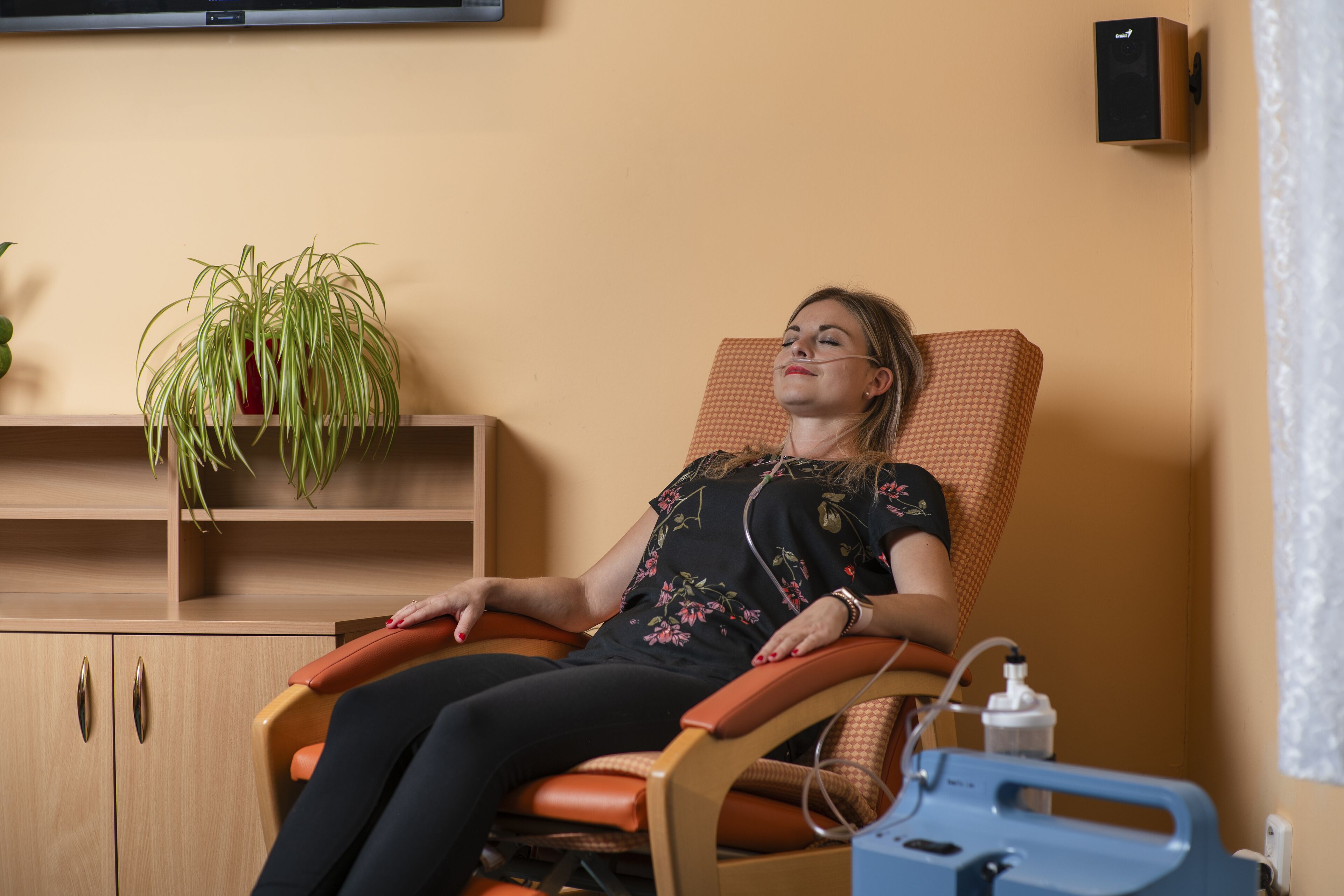 Una persona è sdraiata su una poltrona massaggiante in una stanza con un armadio in legno, una pianta in vaso e un dispositivo medico blu su un tavolo.