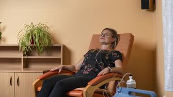 Una persona è sdraiata su una poltrona massaggiante in una stanza con un armadio in legno, una pianta in vaso e un dispositivo medico blu su un tavolo.
