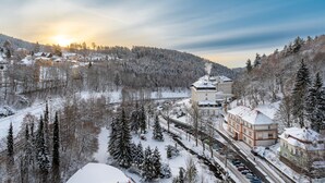 A snow-covered landscape with a building complex, including a large central structure with a chimney emitting smoke, surrounded by snow-laden trees and a road with parked cars. - Hotel Radium Palace (Jachymov)