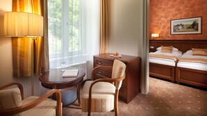 A hotel room with a bed, a desk with a lamp, a chair, a small table, and a window with curtains. - Hotel Radium Palace (Jachymov)