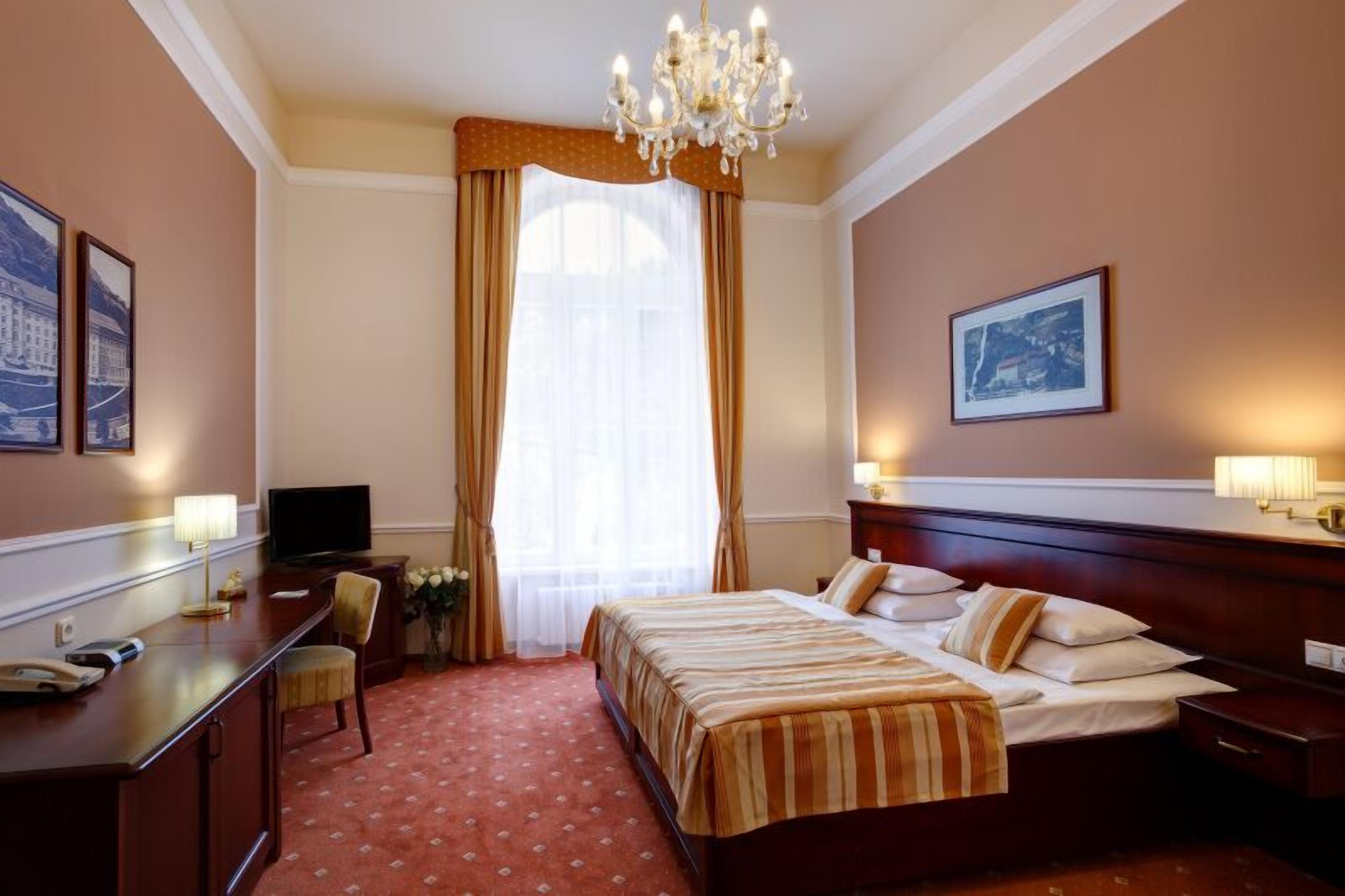 A hotel room with a large bed, a desk with a chair, a television, and a chandelier.