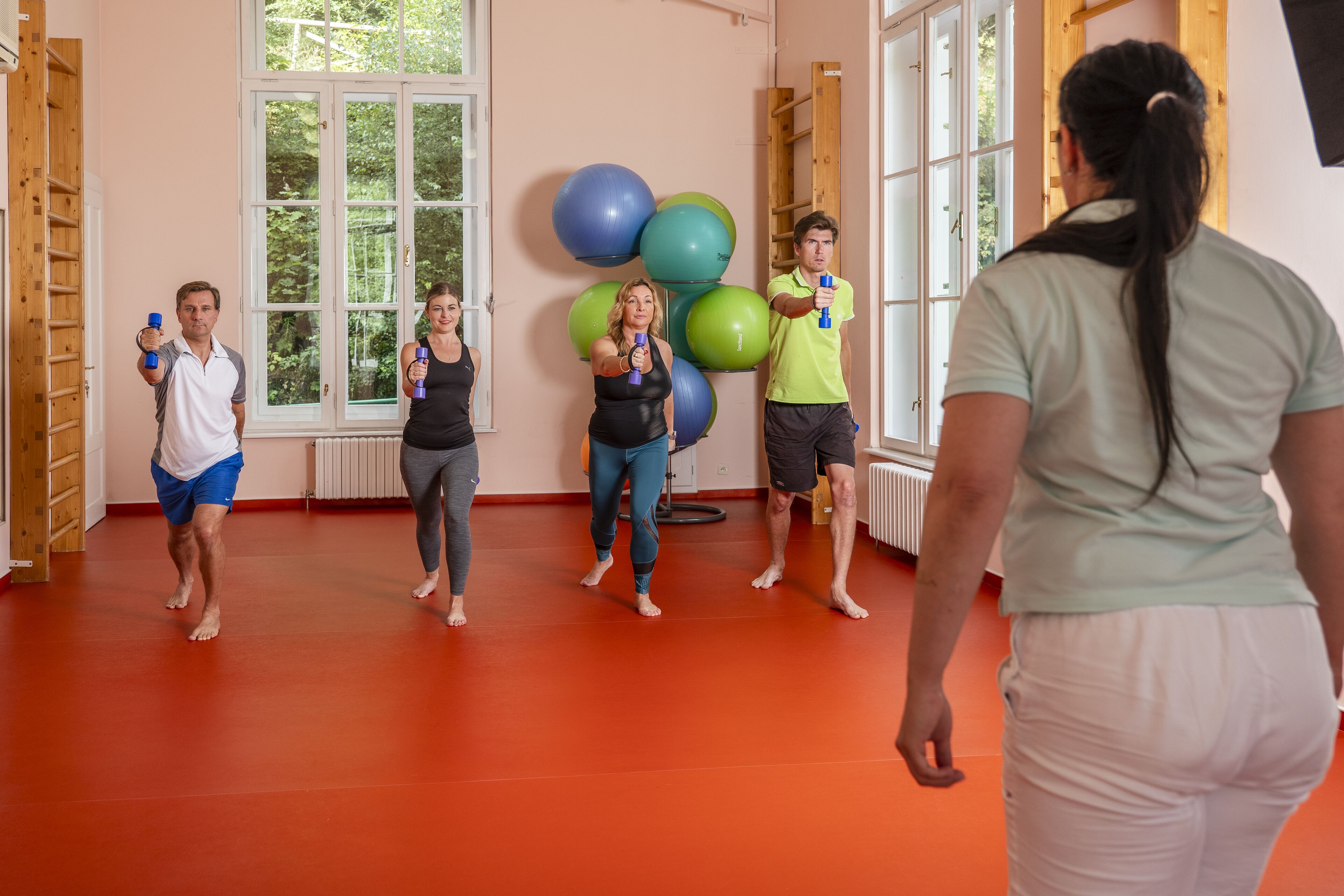 A group of people exercising with dumbbells in a room with large windows, exercise balls, and a red floor.