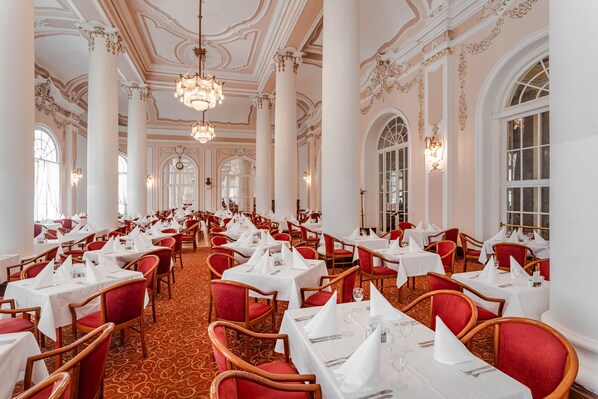 A formal dining room with rows of tables and chairs, large windows, and ornate ceiling details. - Hotel Radium Palace (Jachymov)