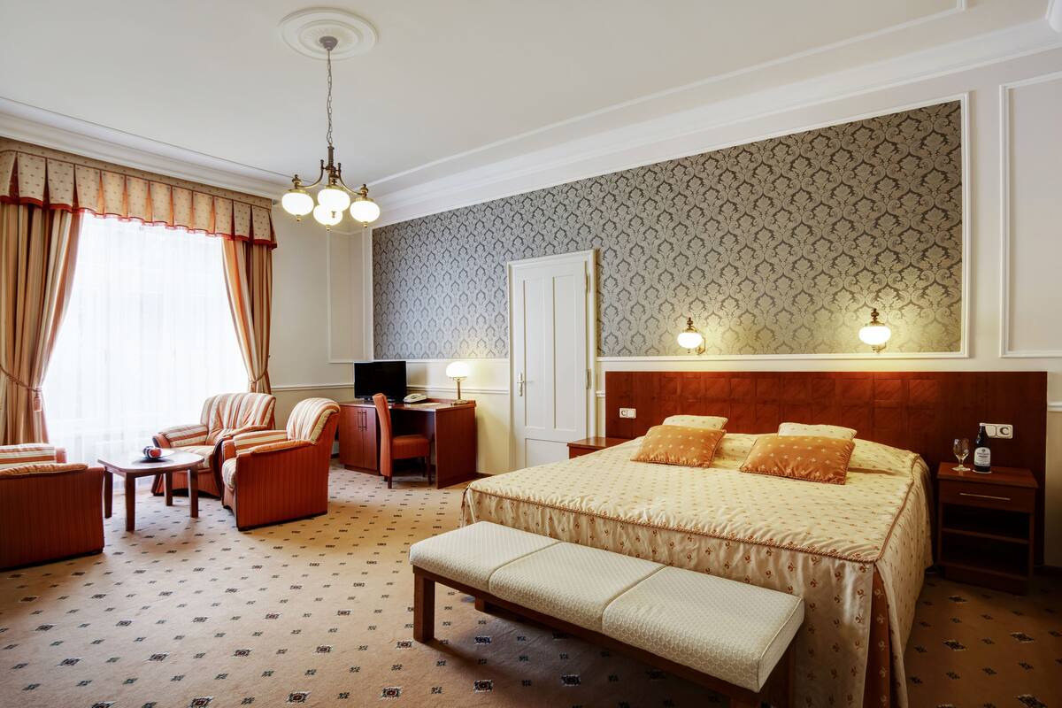 A hotel room with a large bed, a desk with a chair, a small table, a bench, and a TV.