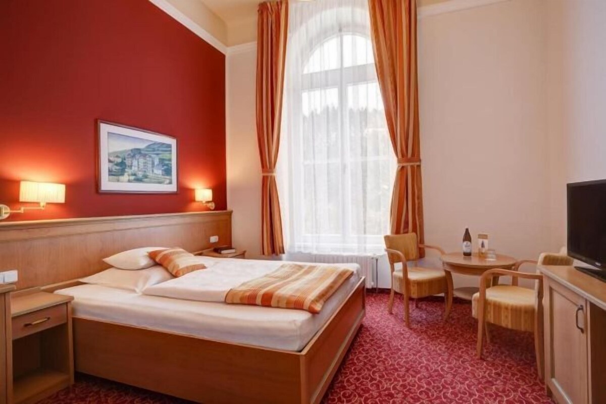 Habitación de hotel con cama, escritorio, silla, televisor y ventana con cortinas.
