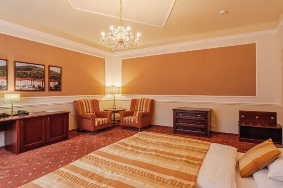 A hotel room with a large bed, two armchairs, a desk with a lamp, and a chandelier.