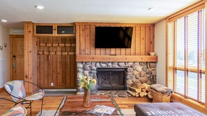 A cozy living room with a stone fireplace, wooden walls, a flat-screen TV, and a large window with blinds. - JHRL - Nez Perce A2 - Beautifully renovated, 1st floor condo (Teton Village)
