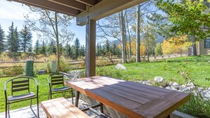 An outdoor patio with a wooden table and two metal chairs, overlooking a grassy area with trees and a mountain in the background. - JHRL - Nez Perce A2 - Beautifully renovated, 1st floor condo (Teton Village)