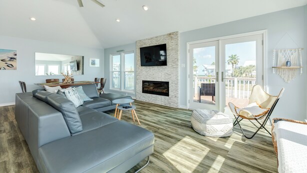 A spacious living room with a grey sectional sofa, a flat-screen TV mounted on a stone wall, a fireplace, and a balcony with a view of palm trees. - Private Beachside Home, Perfect for Families, Updated Interior & Near Beach (Galveston)