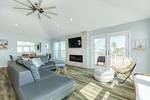 A spacious living room with a grey sectional sofa, a flat-screen TV mounted on a stone wall, a fireplace, and a balcony with a view of palm trees. - Private Beachside Home, Perfect for Families, Updated Interior & Near Beach (Galveston)