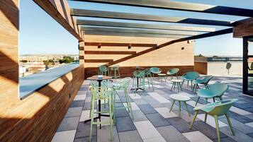 An outdoor terrace with a view of the city, featuring wooden pergola, green and white chairs, and a tiled floor.