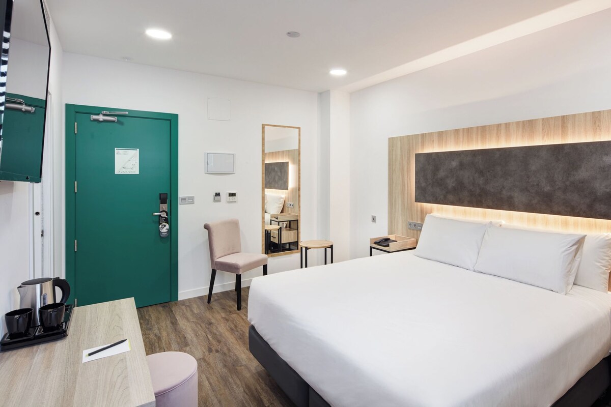 Una habitación de hotel moderna con una cama grande, un escritorio con hervidor, una silla y una puerta verde.