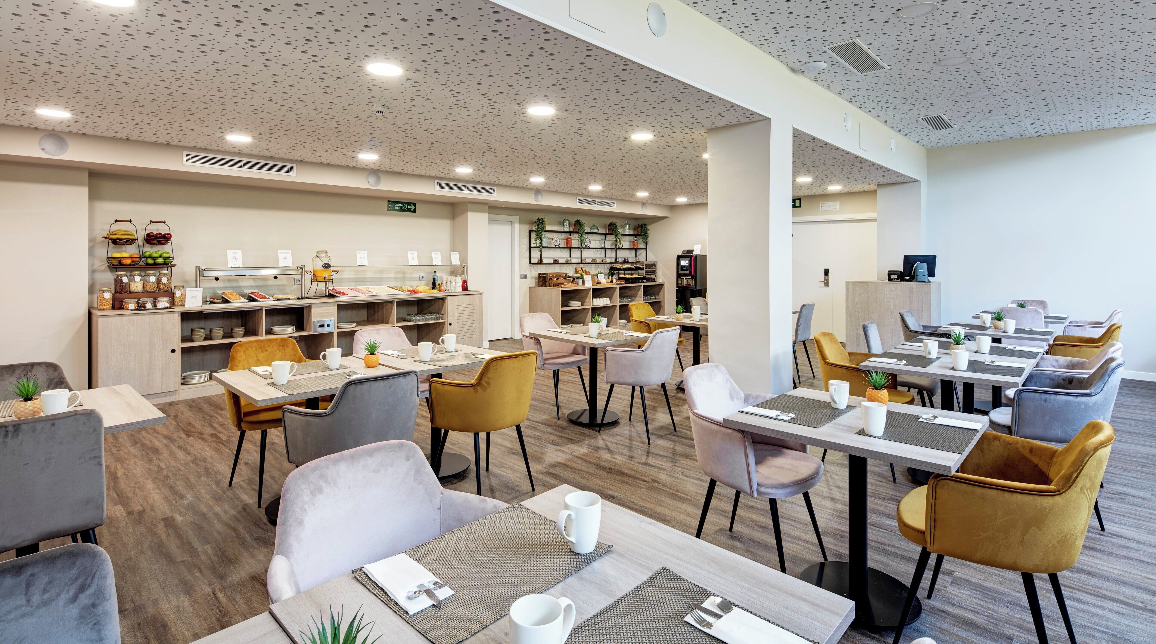 Un comedor moderno con varias mesas y sillas, una zona de cocina con servicio de buffet y un mostrador de recepción.