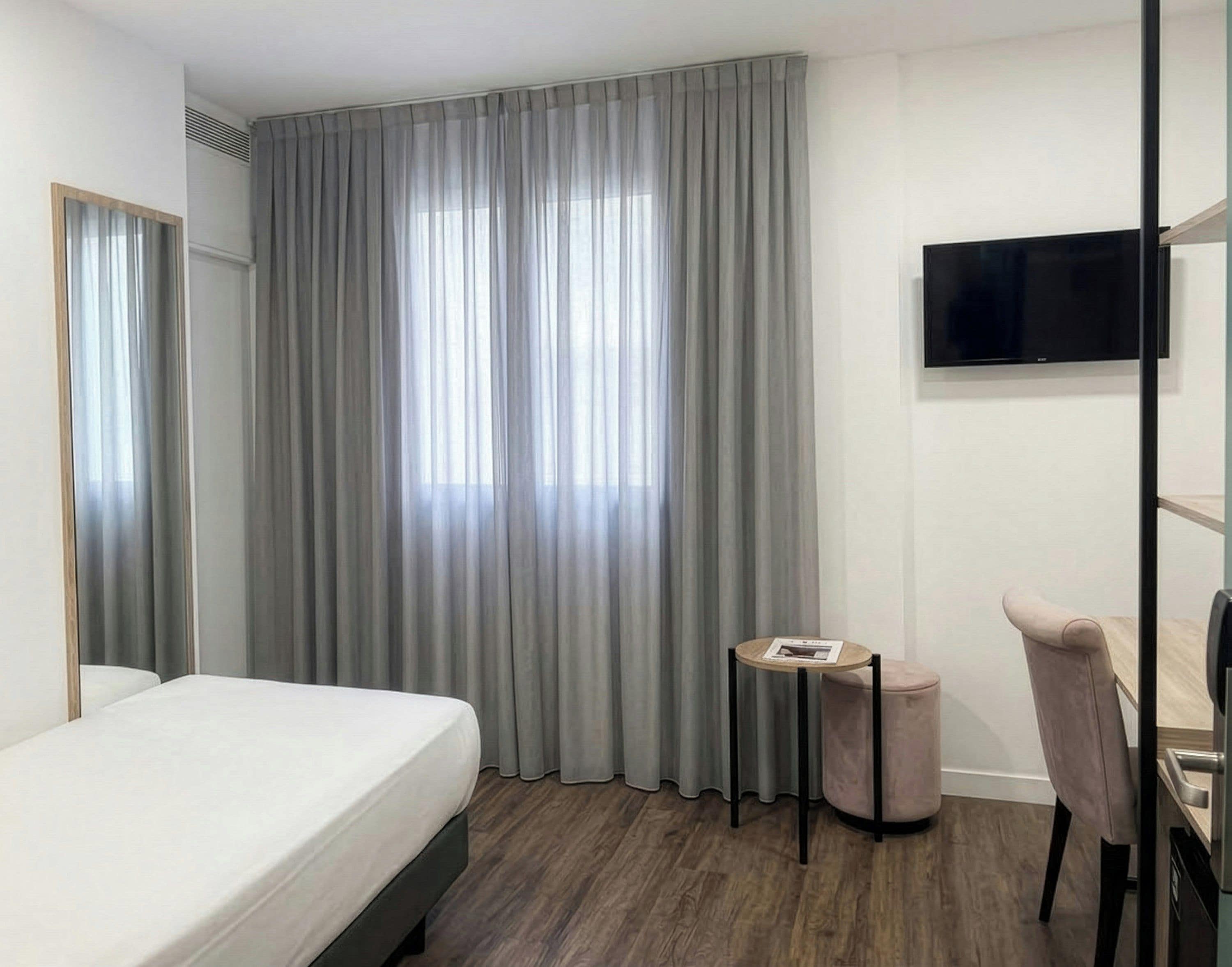 A hotel room with a bed, a small round table, a chair, a television, and a large window with curtains.