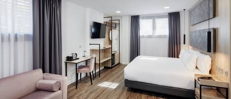 Habitación de hotel moderna con una cama grande, un escritorio con silla, un sofá y un televisor montado en la pared.