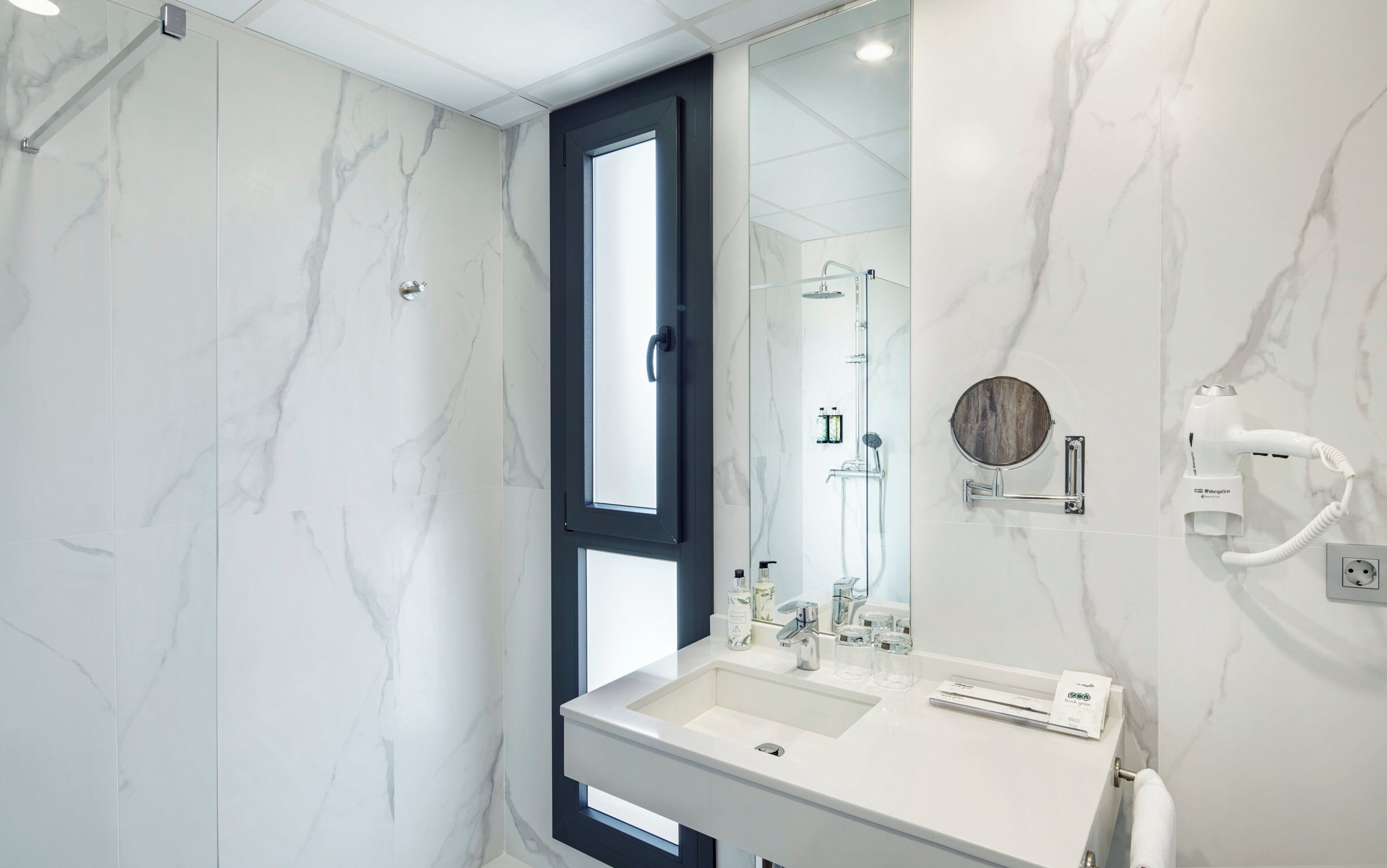 Een moderne badkamer met een marmeren wand, een grote spiegel, een inbouwspoelbak en een aan de muur bevestigde haardroger.