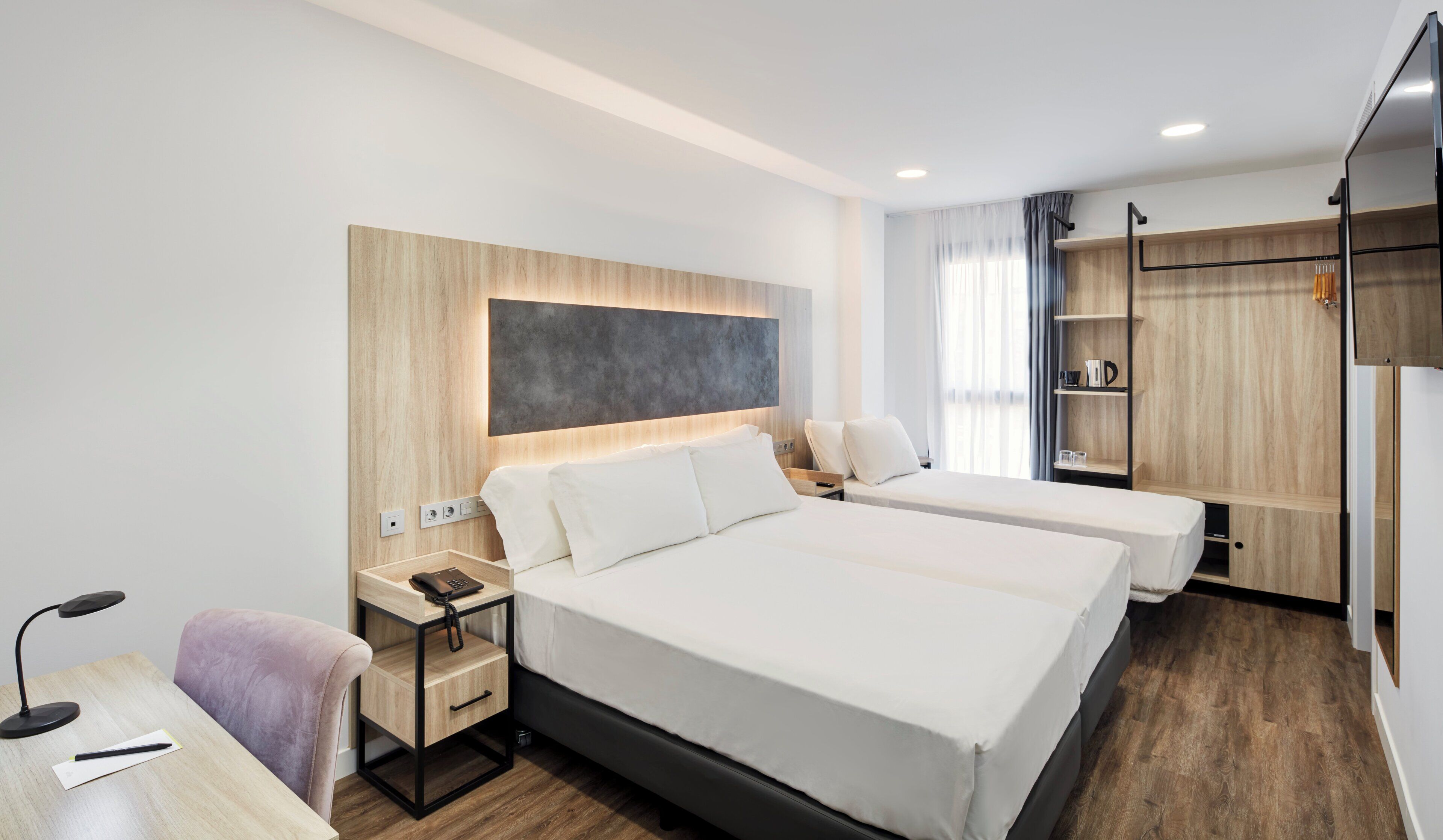 A modern hotel room with two beds, a desk with a lamp, a phone, and a shelf with a coffee maker.