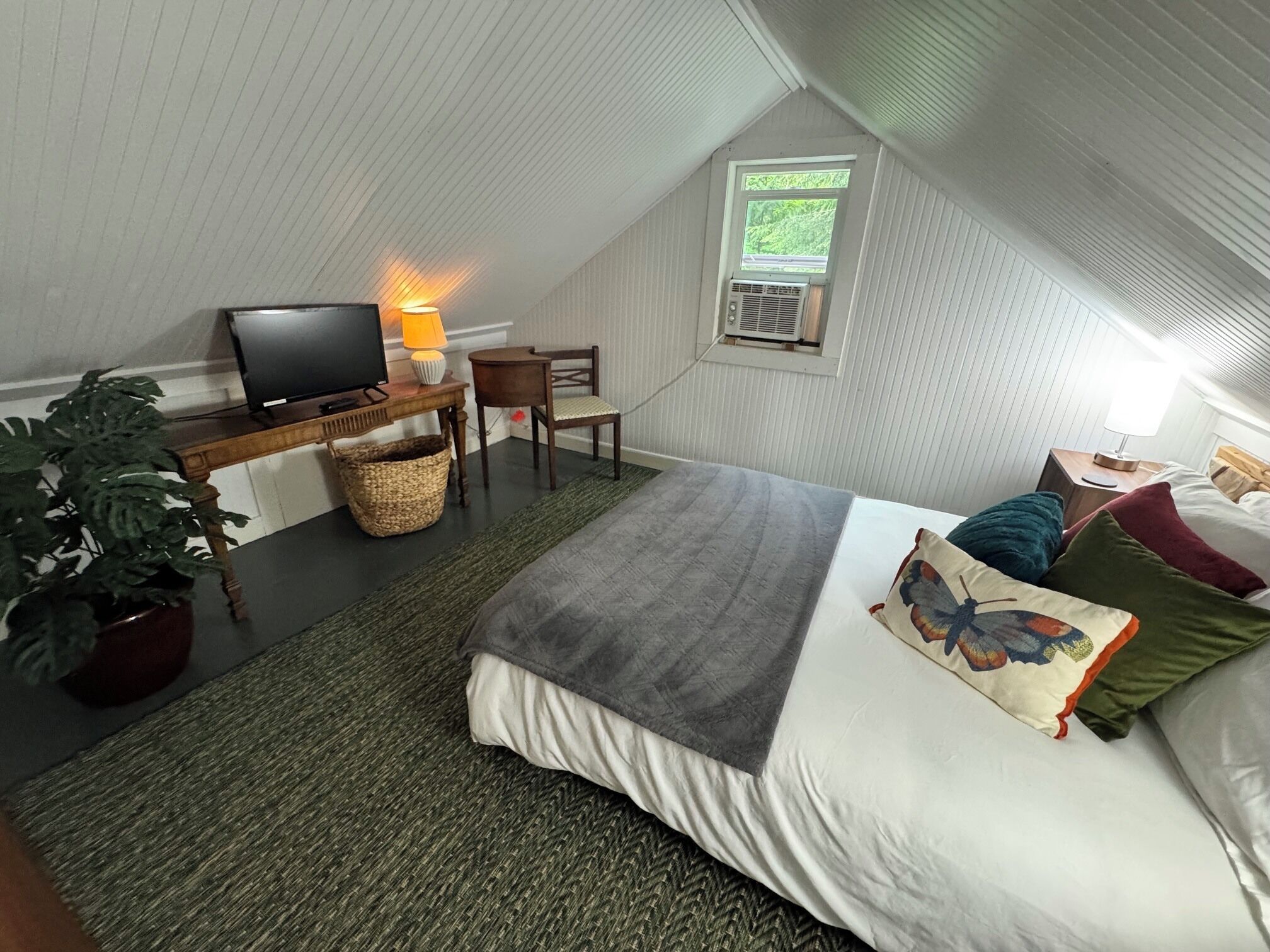 Ein gemütliches Schlafzimmer im Dachgeschoss mit einem Bett, einem Schreibtisch mit Fernseher, einem Sessel, einem Fenster und einer Topfpflanze.