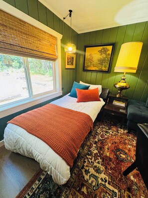 A bedroom with a bed, a window with blinds, a lamp, a rug, and a painting on the wall.